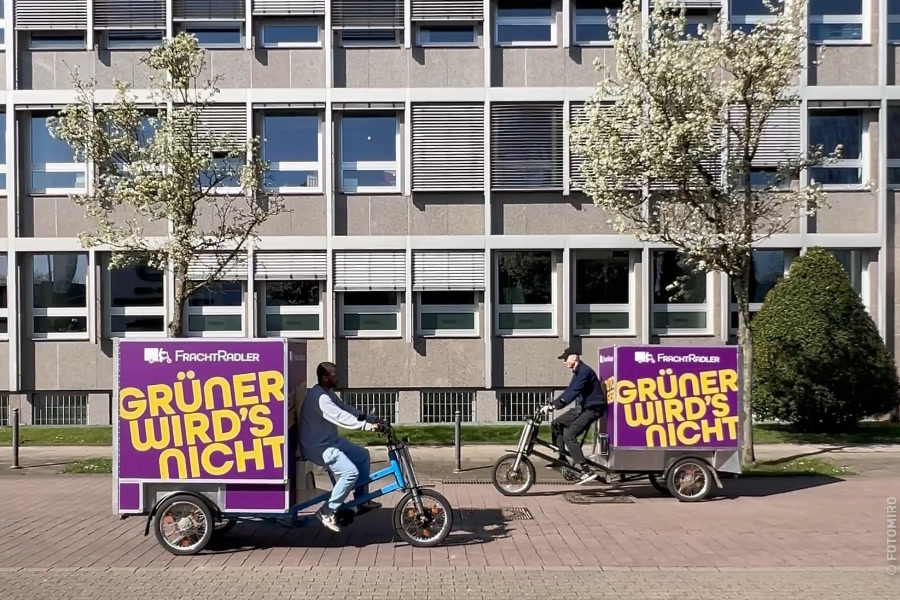 Zwei Männer fahren auf Lastenfahrrädern mit der Aufschrift "GRÜNER WIRD'S NICHT" in der Stadt.